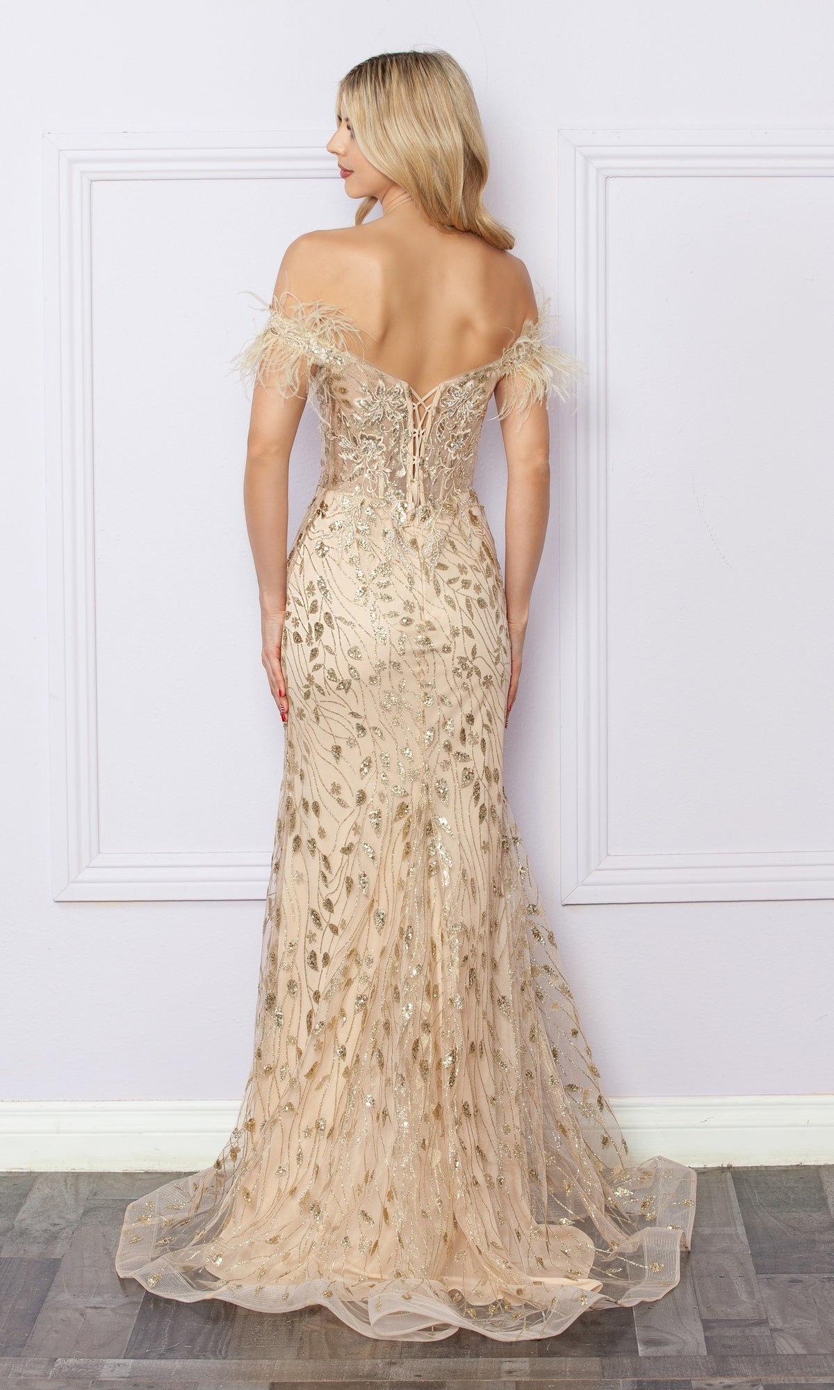 A woman stands facing away, wearing the Nox Anabel L1255 Sequined Glitter-Print Prom Dress in light gold, featuring an off-the-shoulder design, lace-up back, feather-detailed sleeves, and floor-length sequin embellishments.